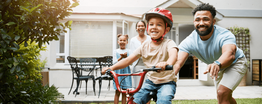 A smiling young child wearing a red helmet rides a bike with help from a man, while a woman and another child cheer in the background outside a home
