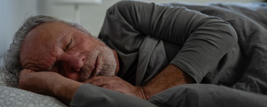 An older man with gray hair and a beard sleeping peacefully in bed, covered with a gray blanket