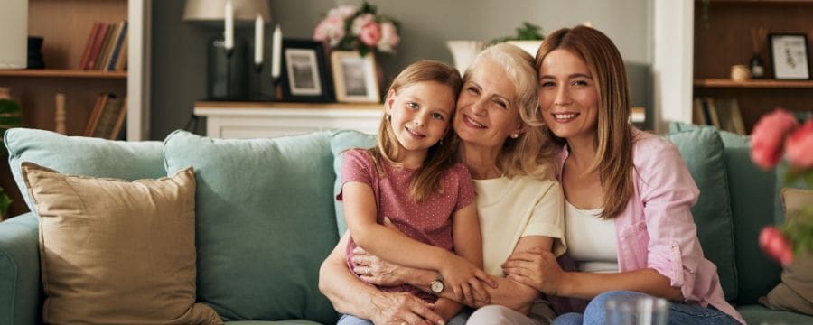 A young girl, an older woman, and a younger woman sitting closely together on a teal couch, smiling warmly at the camera in a cozy living room